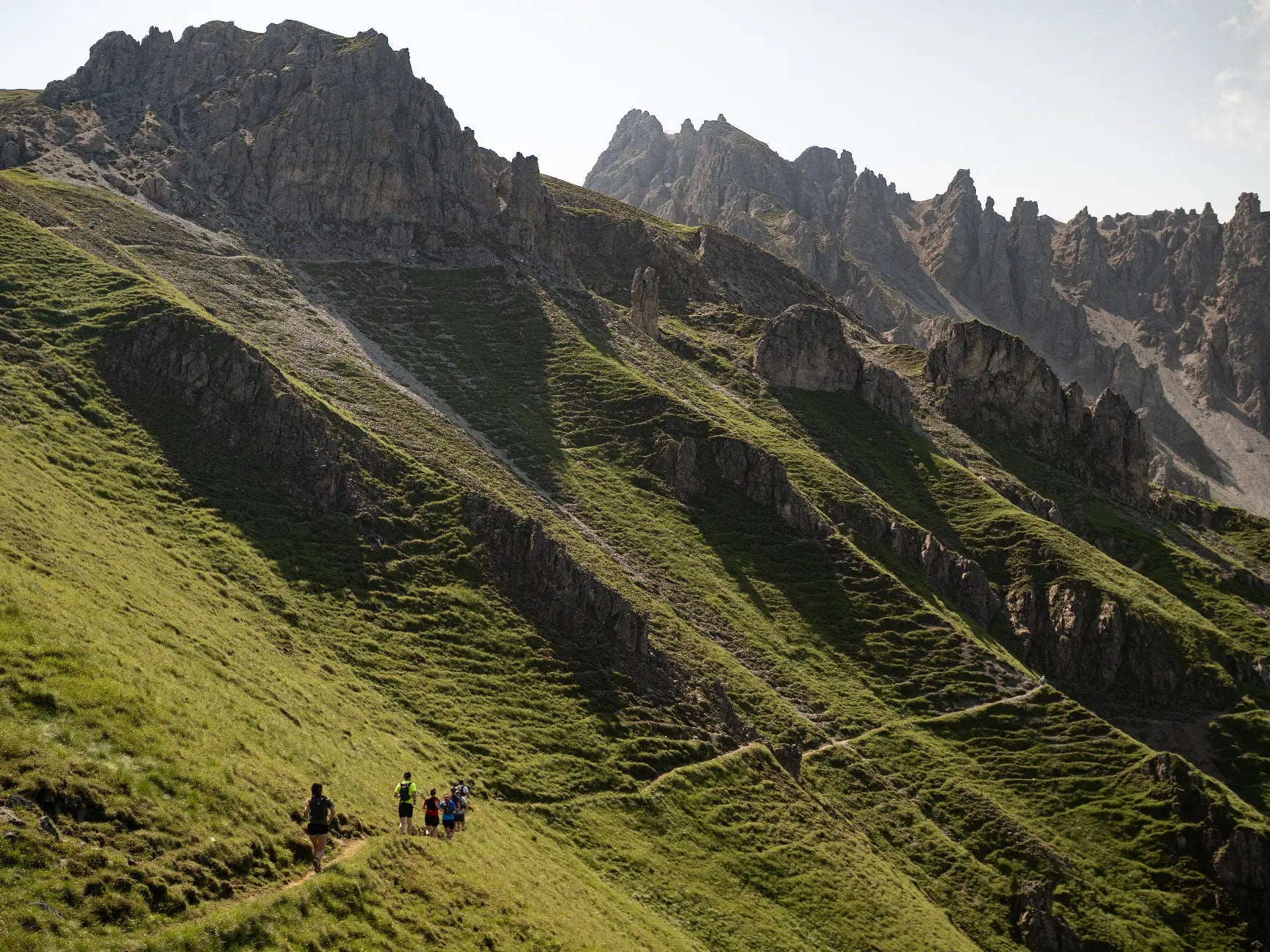 Trail runners on a grassy mountain trail with rocky peaks in the background