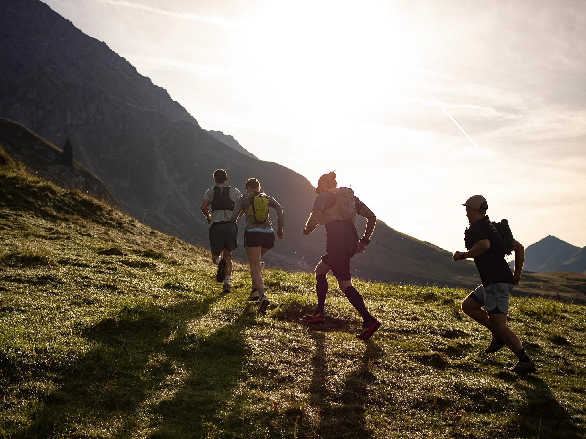 Four people running with trail running vests on a grassy hill with mountains in the background